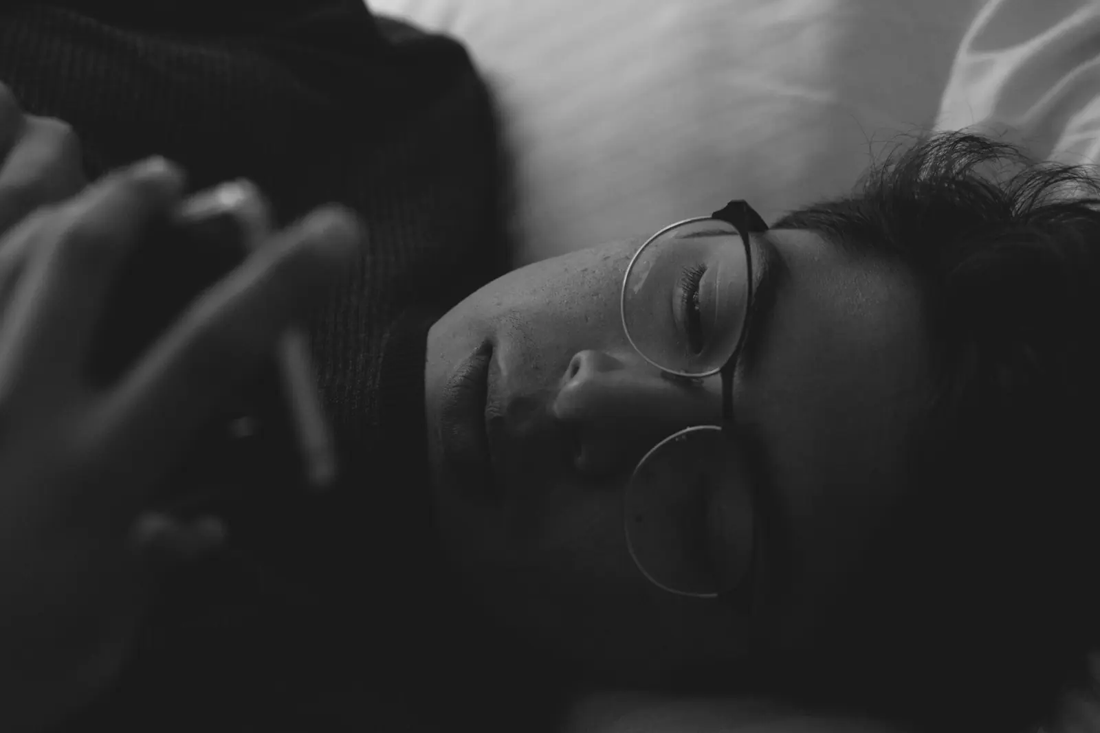 Black and white photo of a young man with glasses lying in bed in a dark room, holding his phone close to his face.