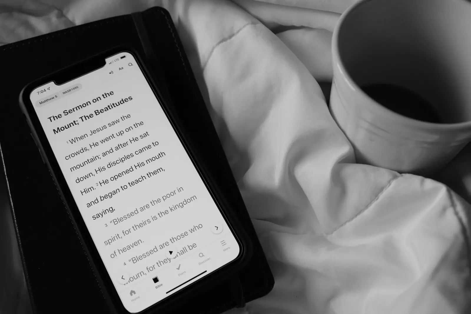 Black and white photo of a phone open to Matthew 5 in a Bible app, lying on a leather notebook next to a mug of coffee on white bedding.